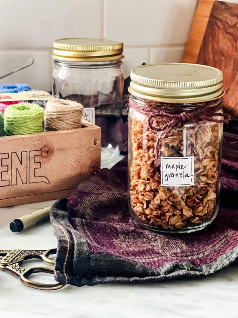 A labeled jar of maple granola with brown twine tied around it sitting on a beautiful towel. In the background a wooden box of colorful twine.