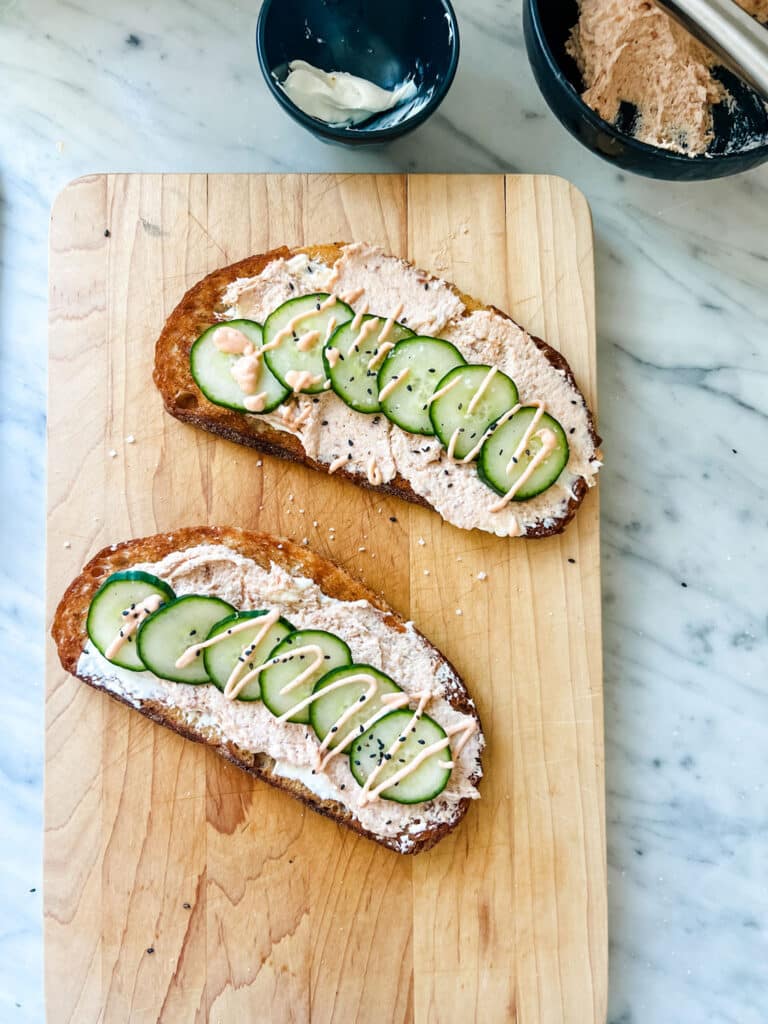 Salmon mousse tartines topped with cucumbers and sriracha mayo sitting on a wood cutting board.