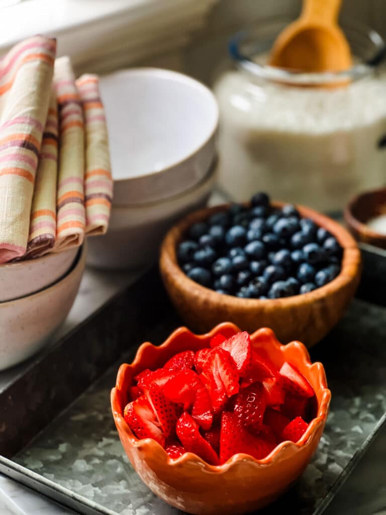 There is an orange bowl of strawberries next to a wooden bowl full of blueberries on a metal serving tray. In the background are bowls for serving, striped napkins, and a jar of shredded coconut.