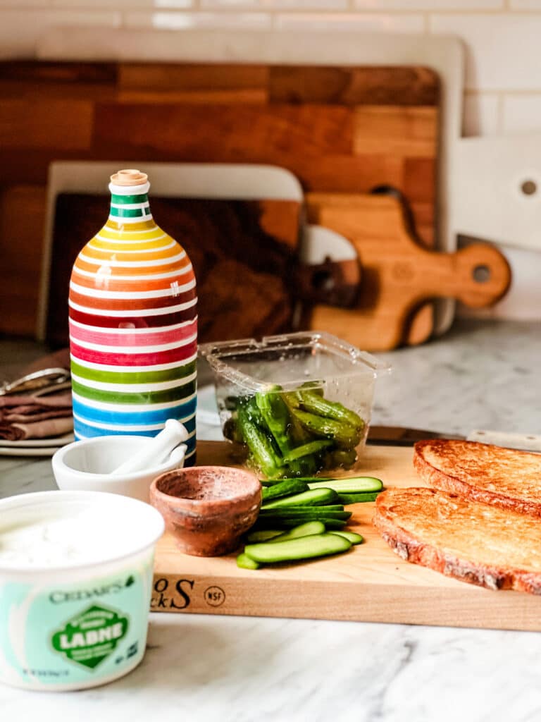 A container of Labneh, cucumbers, striped olive oil bottle on cutting board