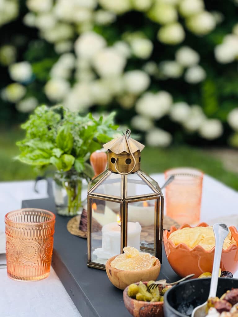 Lantern with lit candle, fresh herbs, and cheese and charcuterie board with orange drinking glasses in background