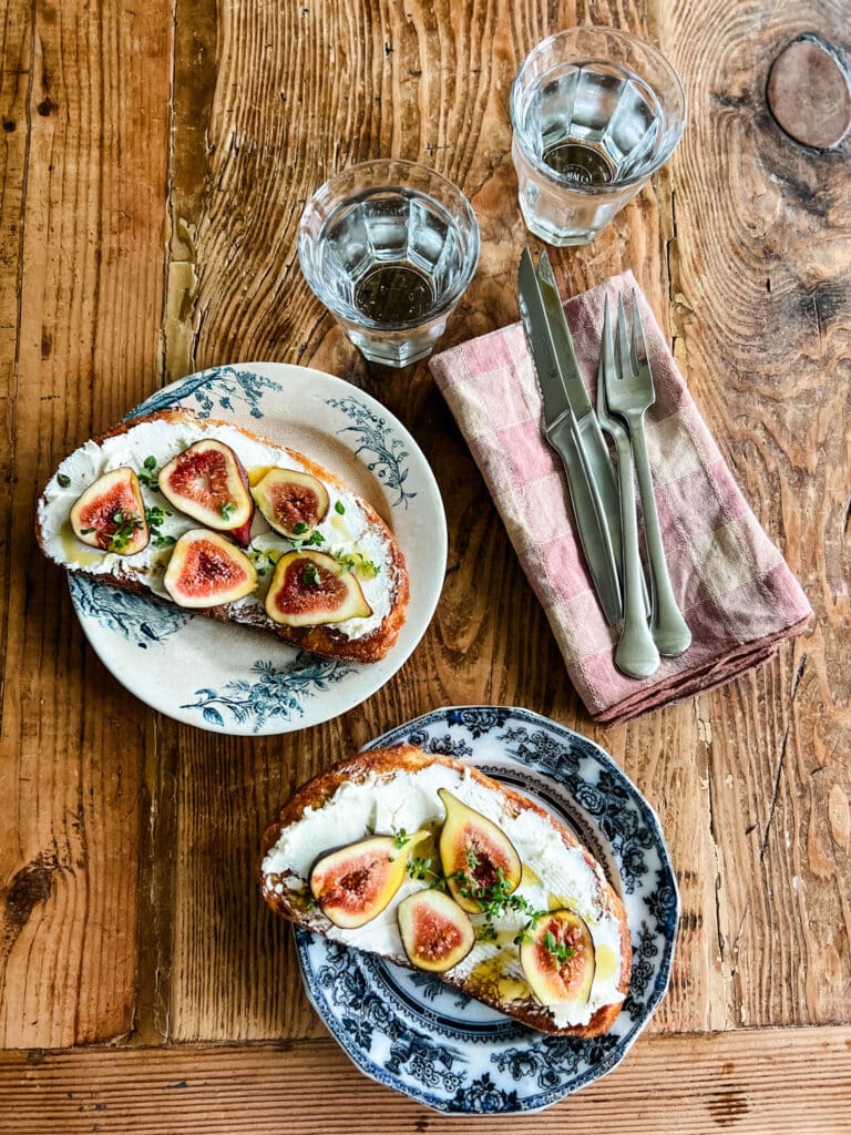 Two fresh fig and goat cheese tartines on patterned plates sitting on a wooden dining table with two glasses and silverware sitting on napkins 