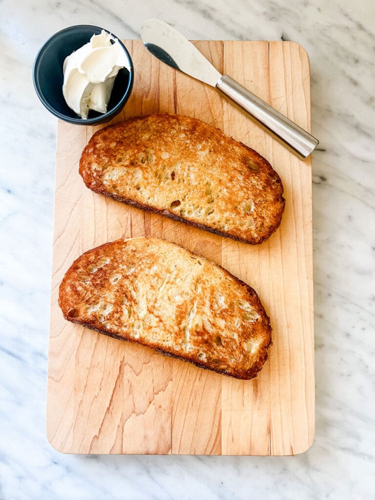 Toasted Italian bread on a wood cutting board with cream cheese and a spreader.