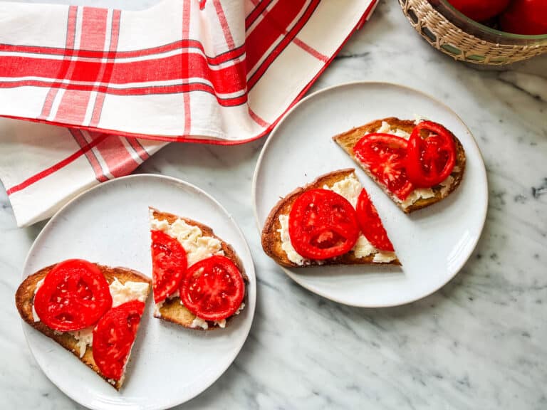 Fresh tomatoes with melted cheddar mayo on buttered toast is served on small white plates and a French tea towel is on the counter next to the white plates.