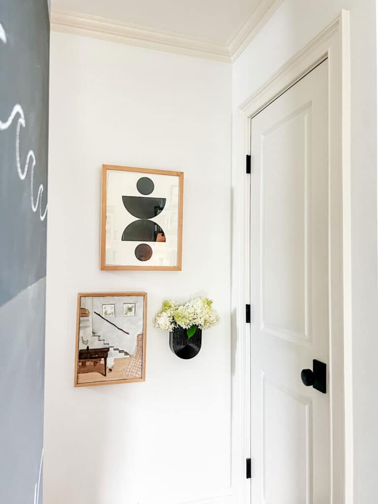 Three pieces of art hanging on a wall. One customer piece from Framebride, a small oiling paint of a staircase and a black wall vase with fresh cut flowers.