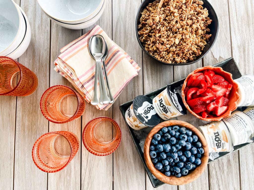 Maple granola in a black bowl, a tray with fresh strawberries and blueberries on a tray with Noosa yogurt. Sitting on a teak table with white bowls, spoons on top of napkins and orange glasses.