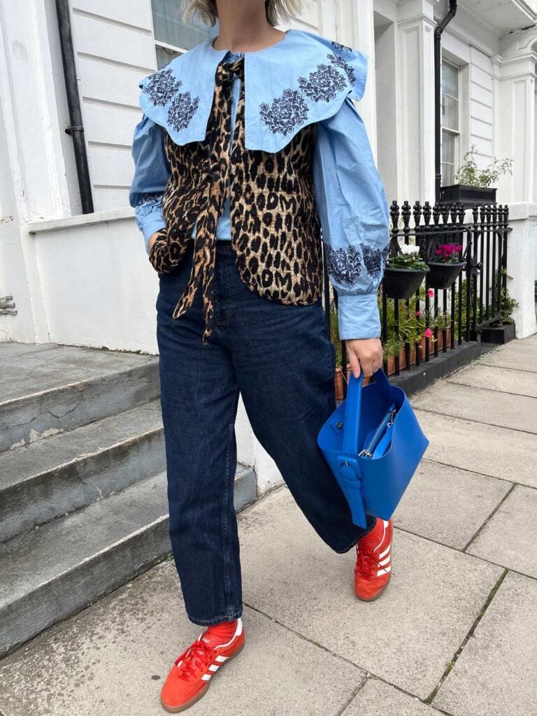 A woman walking is wearing a leopard vest, jeans and red Adidas Gazelles.