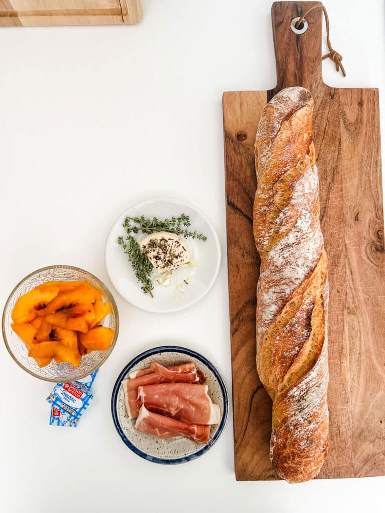 A crusty French baguette on a wood cutting board sitting next to some savory ham, fresh sliced summer peaches and locally sources goat cheese with thyme springs.