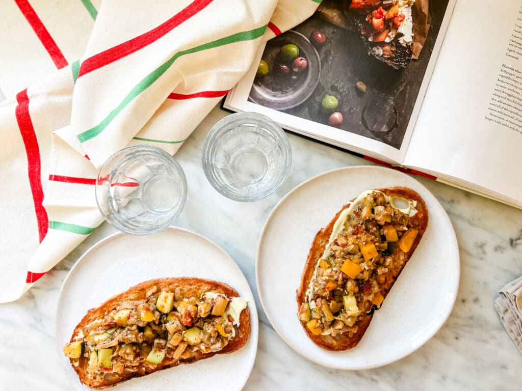 Two white plates with ratatouille tartines sit on a marble countertop with two clear glasses, a vintage French tea towel, and the Buvette cookbook.