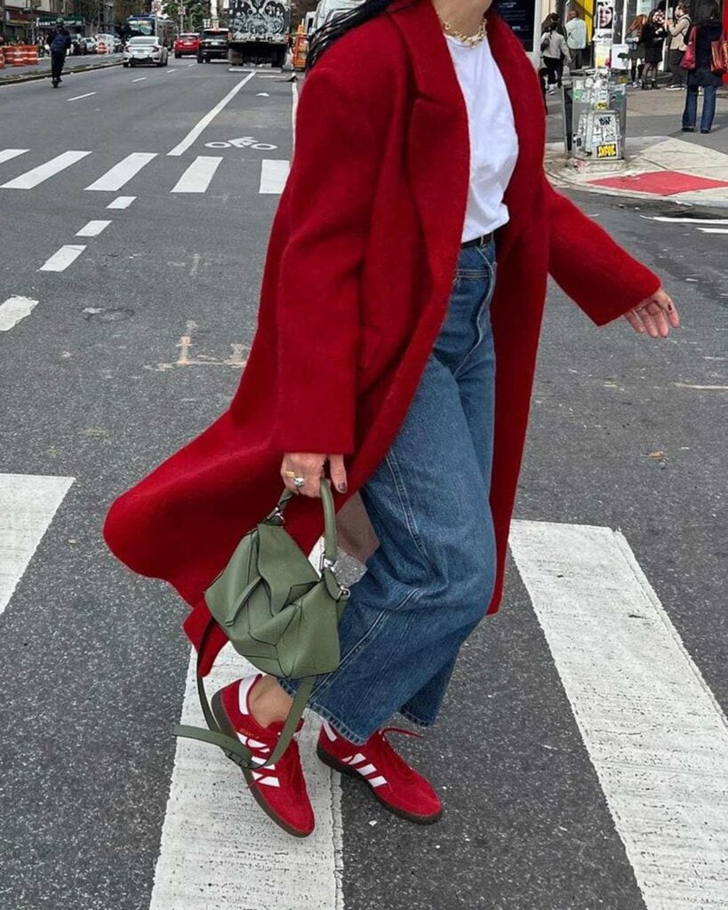 A woman crossing the street wearing a red coat and red Adidas Gazelles.