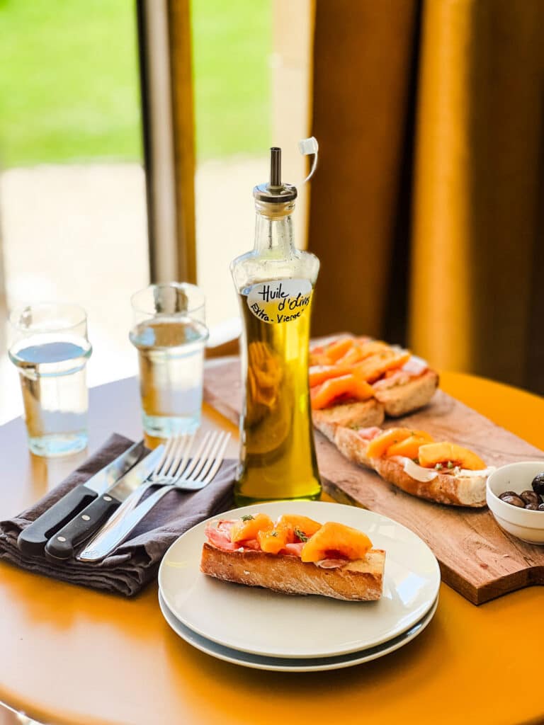 A tartine is served on a white plate in the foreground. In the background is silverware, two glasses of sparking water, a bottle of olive oil, and a cutting board with more tartines.