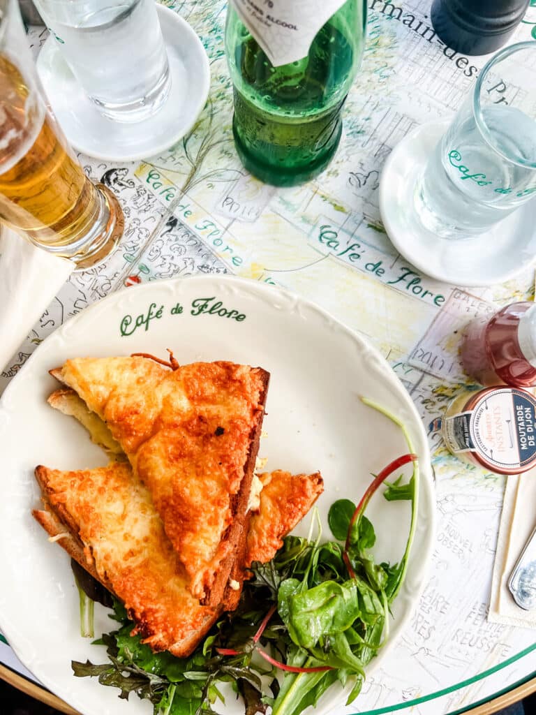 A traditional taste of Paris is the Croque Monsieur and it sits on a white plate with a stenciled logo of Cafe de Flore on it. It is served with a small green salad.