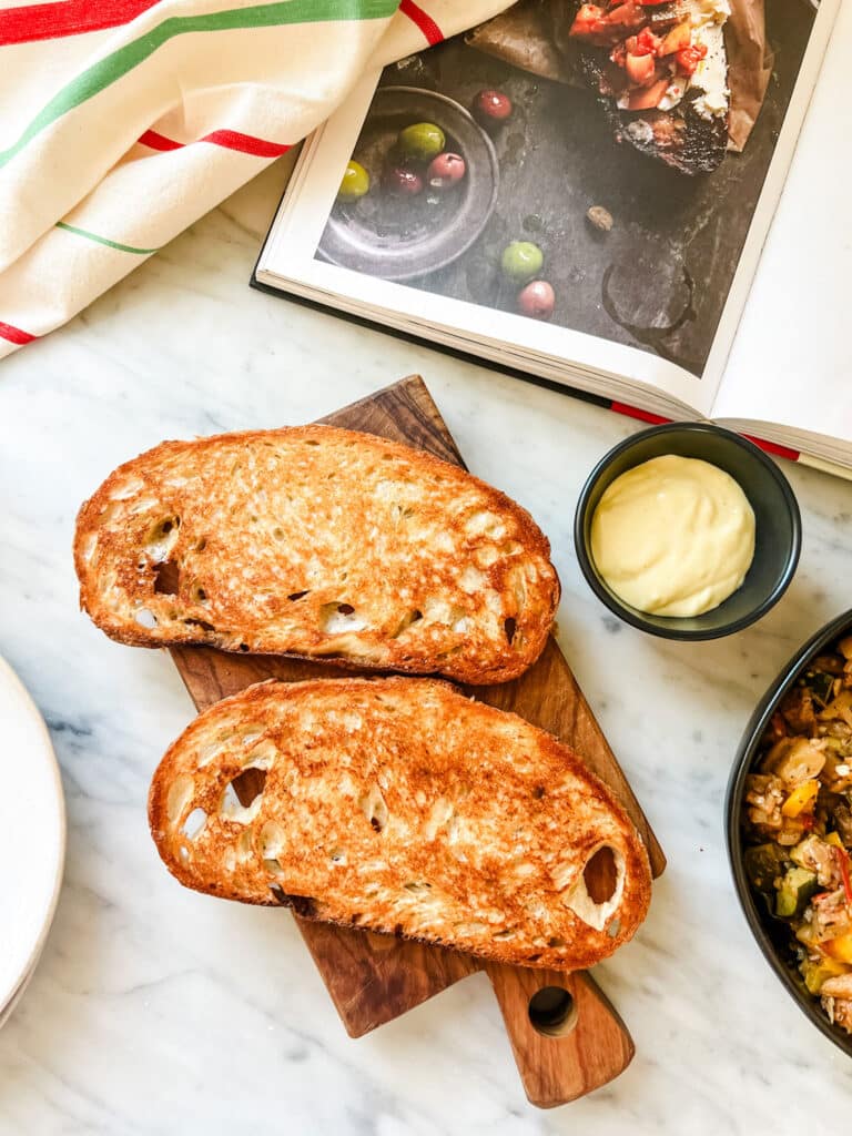 Two slices of grilled bread are resting on a wood Italian cutting board next to a homemade aioli and a bowl of cooked vegetables.