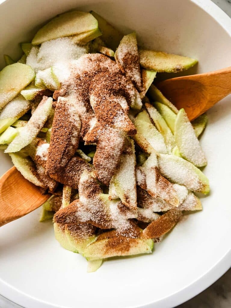 Slices apples with cinnamon and sugar are in a white mixing bowl ready to be tossed with wooden spoons.