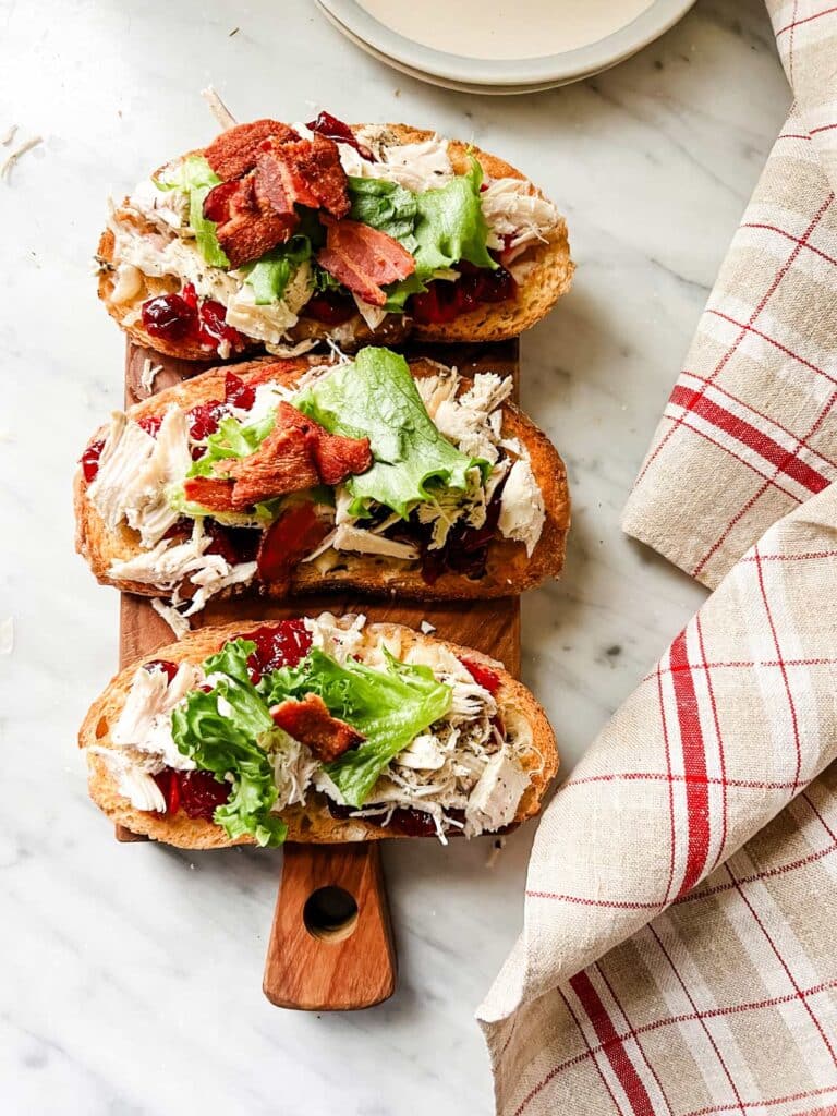 Three roasted turkey tartines are topped with lettuce and bacon and are sitting on a wooden cutting board.