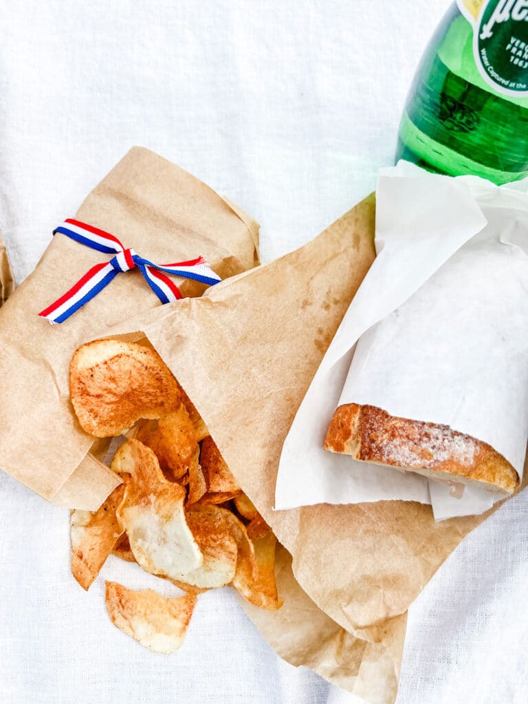 Homemade potato chips are in paper bags and sitting next to a sandwich wrapped in butcher paper.