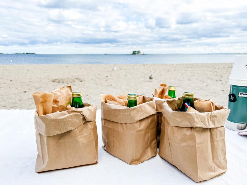 Paper bags filled with lunch and a drink are sitting on a tablecloth draped over a picnic table with the sand and ocean in the background.