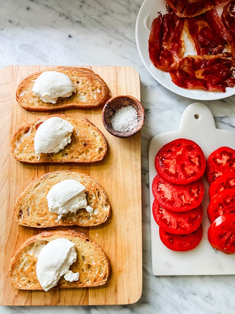 Toasted bread is topped with fresh burrata and is waiting for the sliced tomatoes and fried prosciutto.