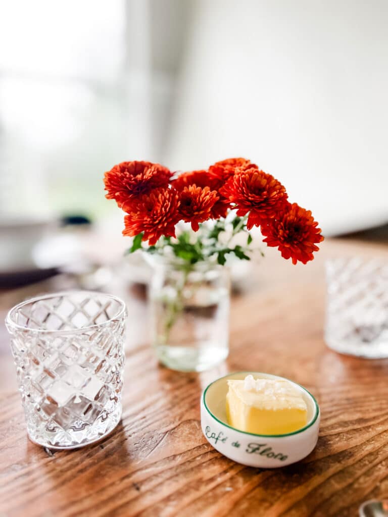 Small red/orange flowers, two small glass votives and butter in a small Cafe de Flore dish decorate and dining room table.