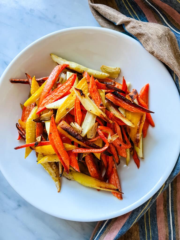 Maple-glazed autumn carrots with sliced garlic cloves are roasted and served in a white bowl.
