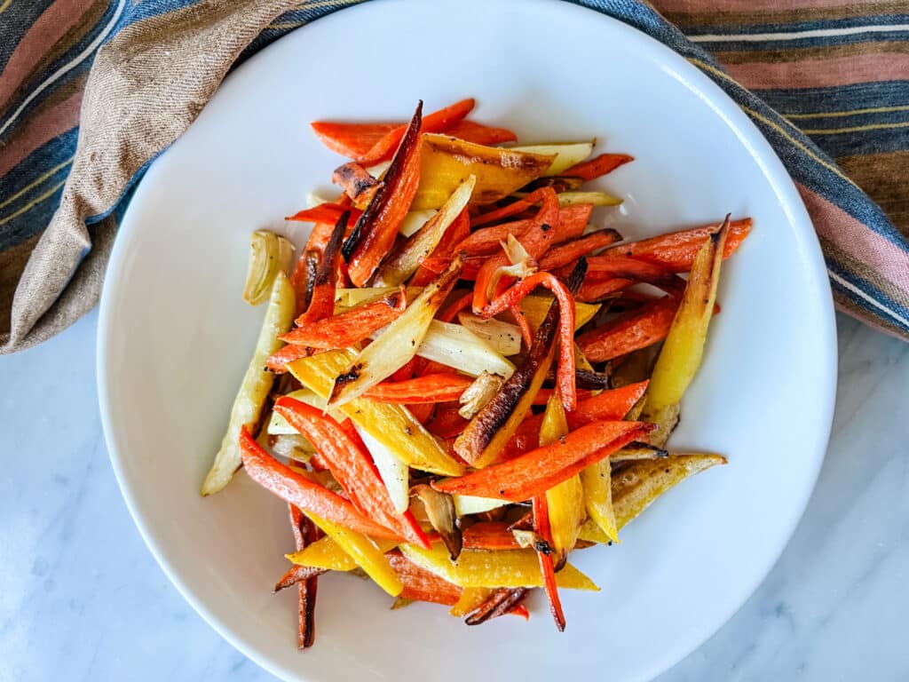 Maple-glazed carrots are in a white serving bowl. Alongside is a brown and white Belgian tea towel.