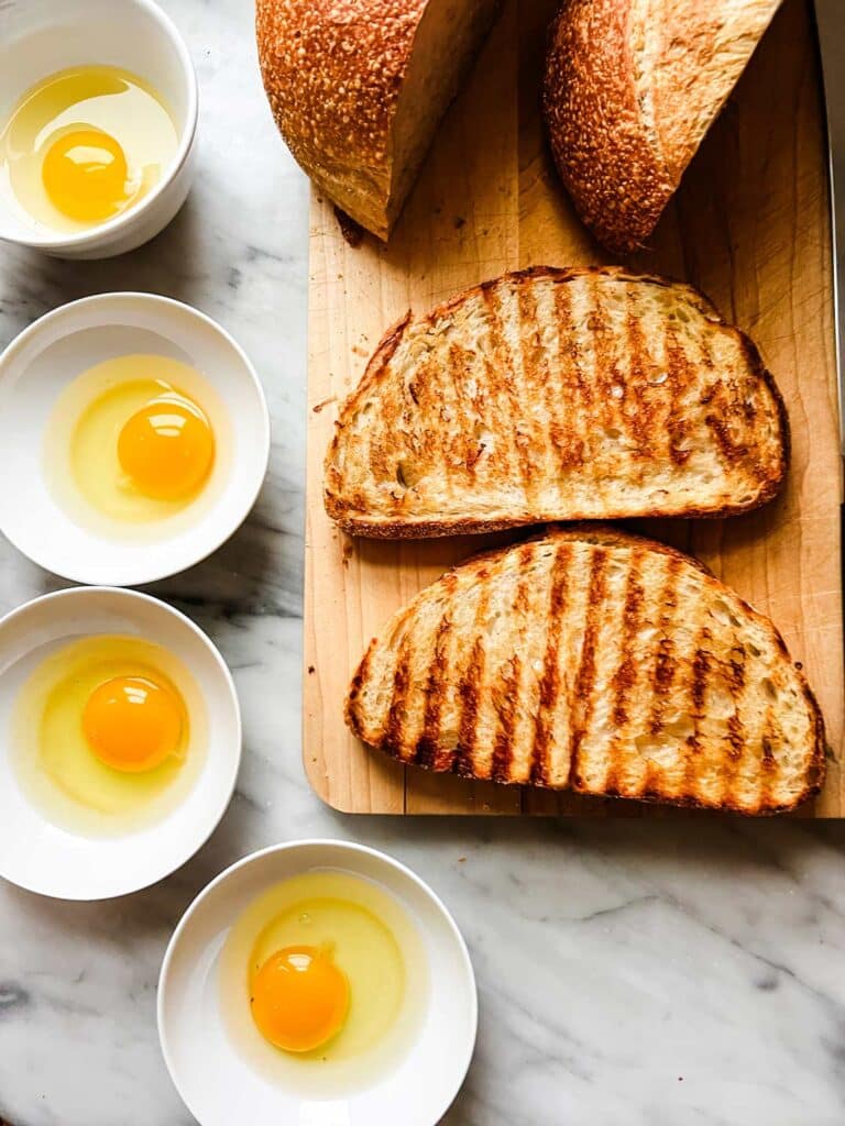 The bread has been toasted on a grill. Next to the bread are four small white bowls each with an egg cracked in it.