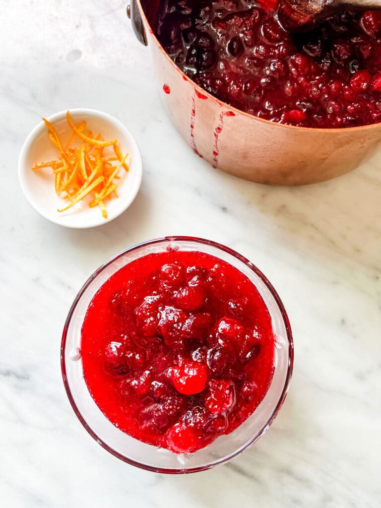 Glass jar of cranberry sauce, small white bowl of orange zest and a copper  saucepan with cranberry sauce.