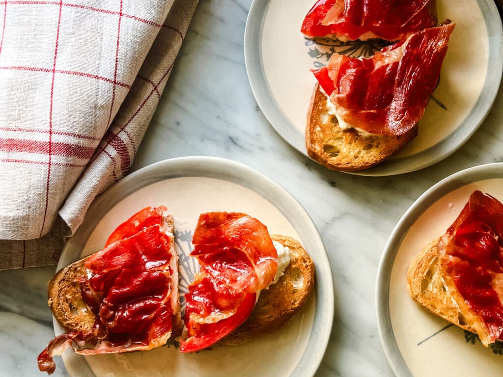 Burrata, tomato and prosciutto tartines are a very nice way to use the last of your summer tomatoes. They are served on small blue and white plates withe a French tea towel next to them.
