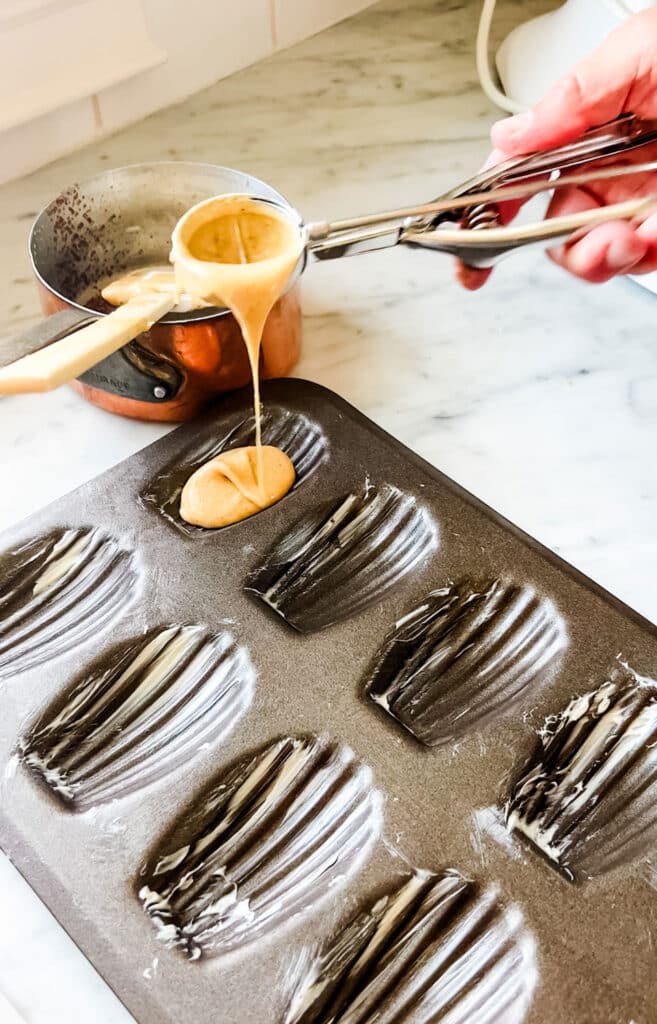 A Madeleine tin sits on a marble countertop and is being filled with batter to pop into the oven.