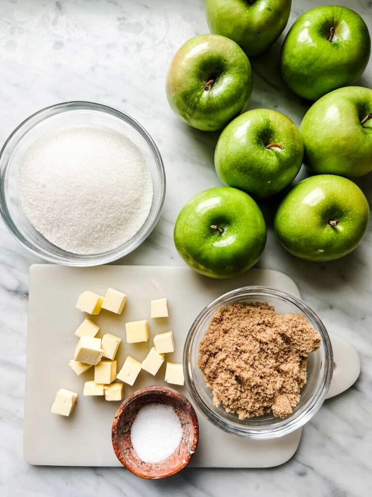 Seven granny smith apples are sitting next to a bowl of white sugar, brown sugar, cubes of cold butter and salt in a small bowl.