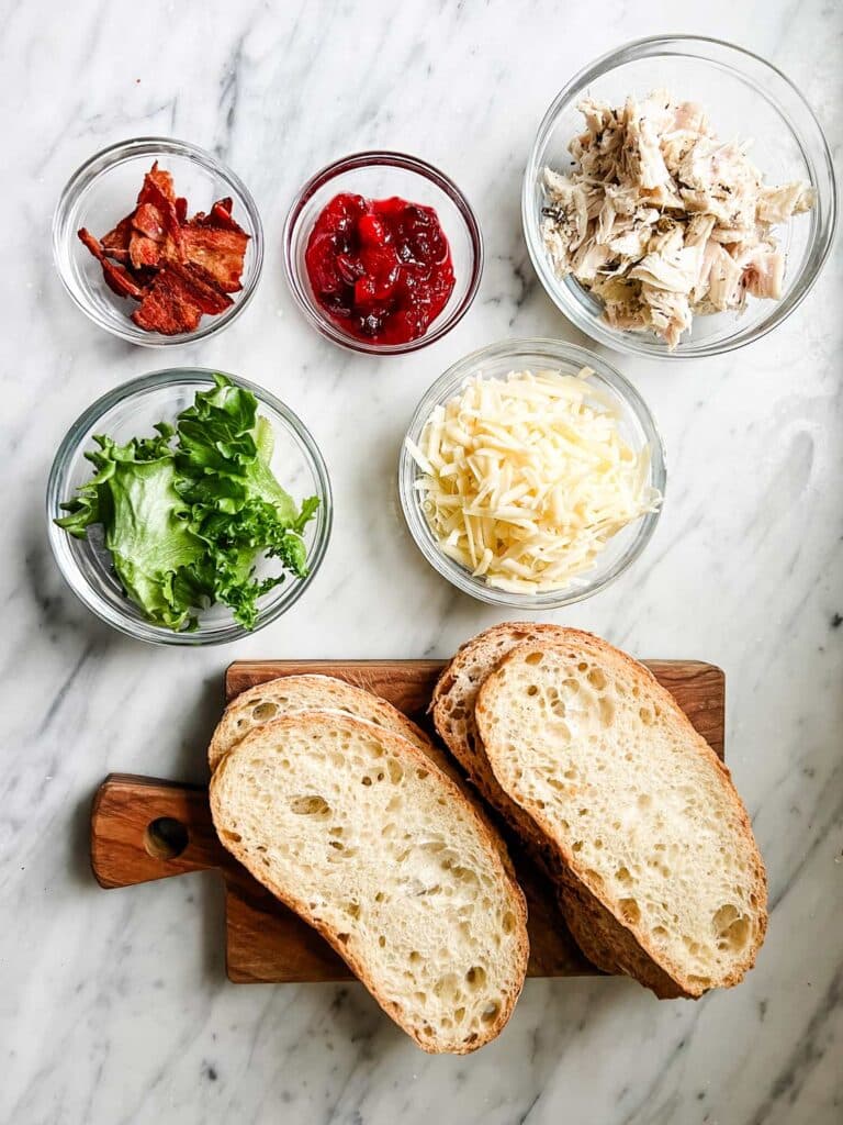 In small glass bowls are bacon, cranberry sauce, turkey, lettuce and cheese. Next to them are four slices of country bread on wooden cutting board.