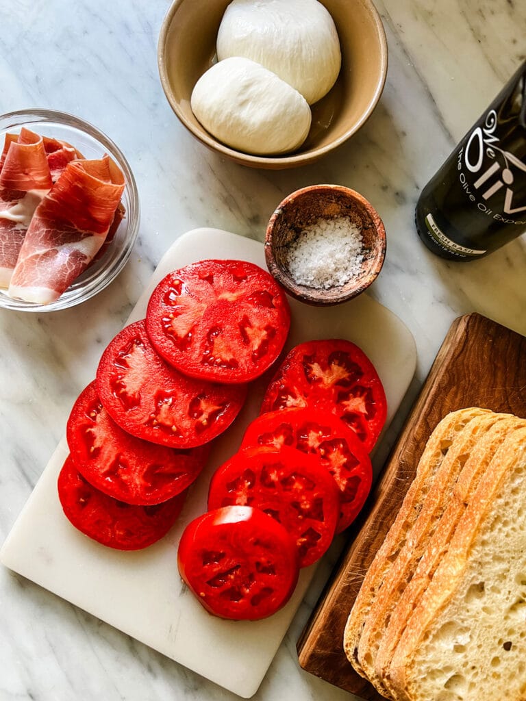 Sliced tomatoes and sea salt rest on a marble cutting board. They are surrounded by bowls of prosciutto and burrata. Next to them on a wood cutting board are slices of Italian country bread.