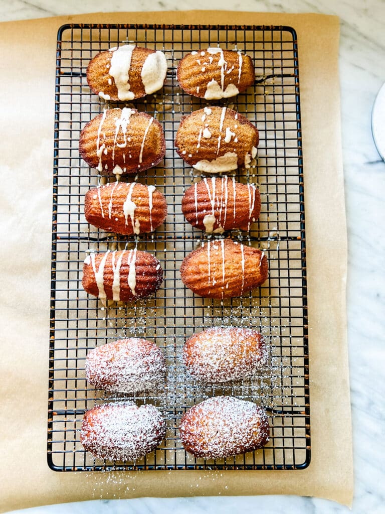 Fresh Madeleines, both glazed and powdered with sugar, rest on a cooling rack.