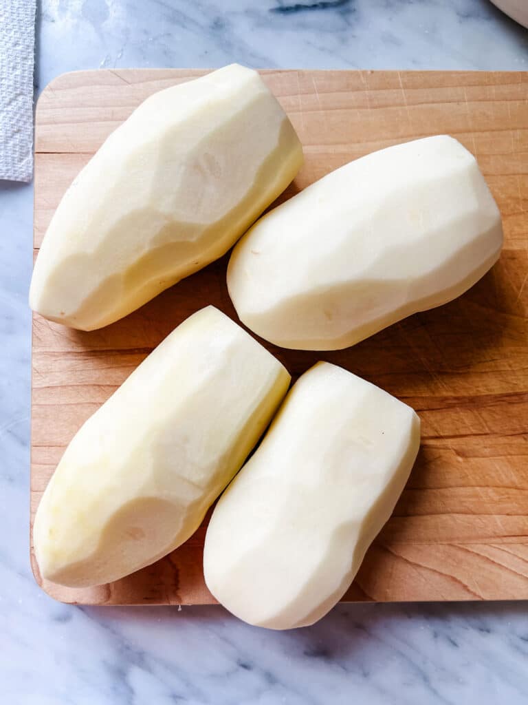 Four peeled potatoes are sitting on a wooden cutting waiting to be sliced.