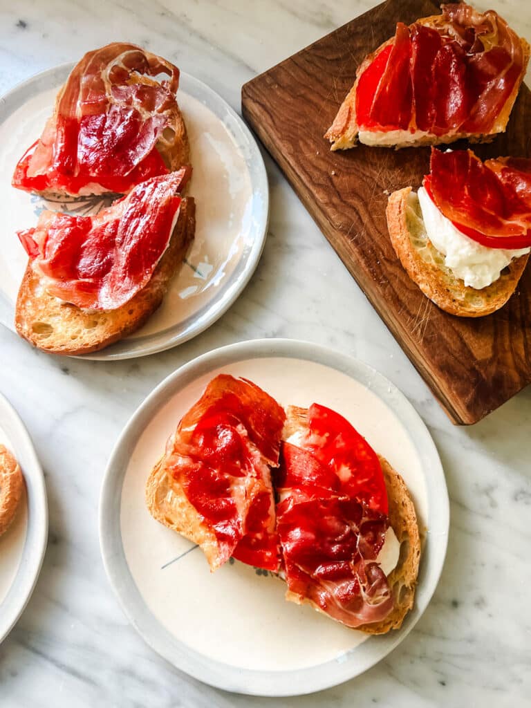 Burrata, tomato, and fried prosciutto tartine are served on small white plates with light blue rims. A few of the tartines are on a wooden cutting board.