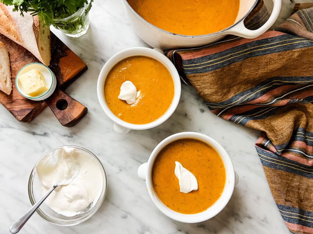 A pot of freshly made pumpkin soup is in a dutch oven. In the foreground are two white bowls, full of soup, with a dollop of creme fraische. A baguette with butter is on a wooden cutting board.