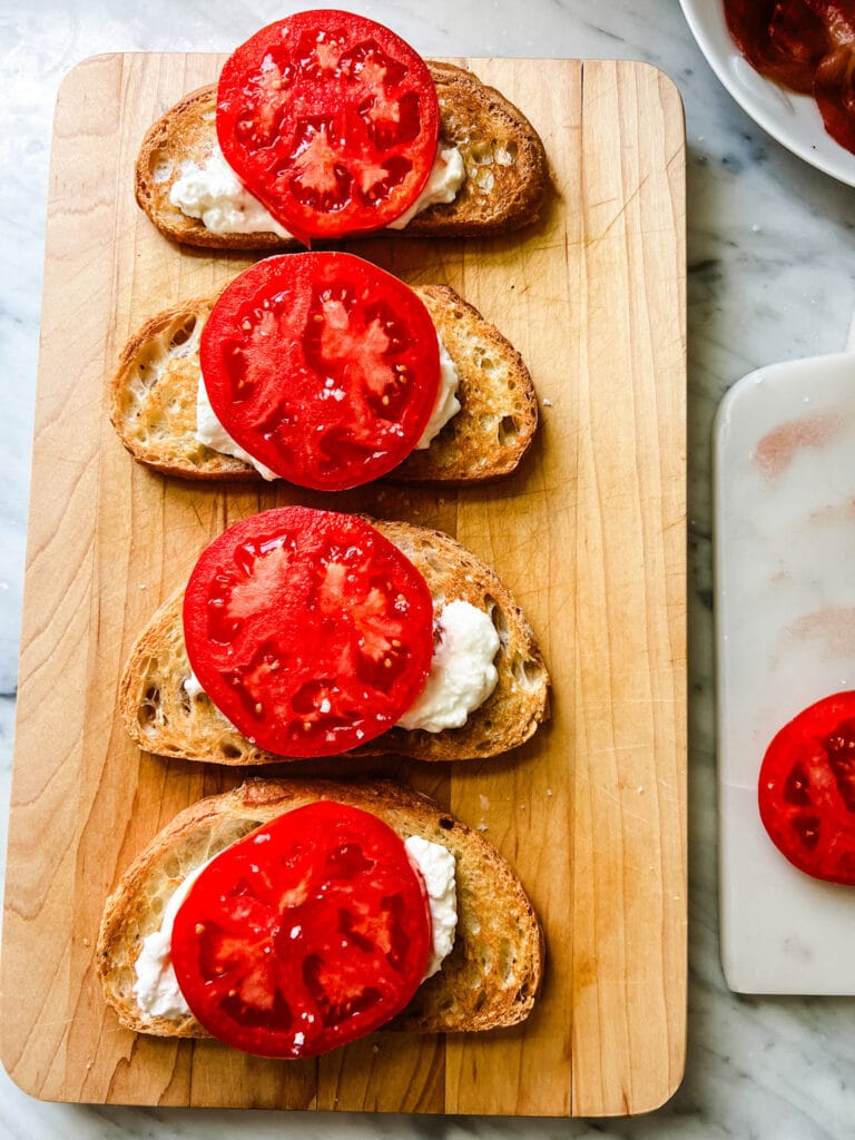Toasted bread is sitting on a wooden cutting board and is topped with fresh burrata and sliced tomatoes.