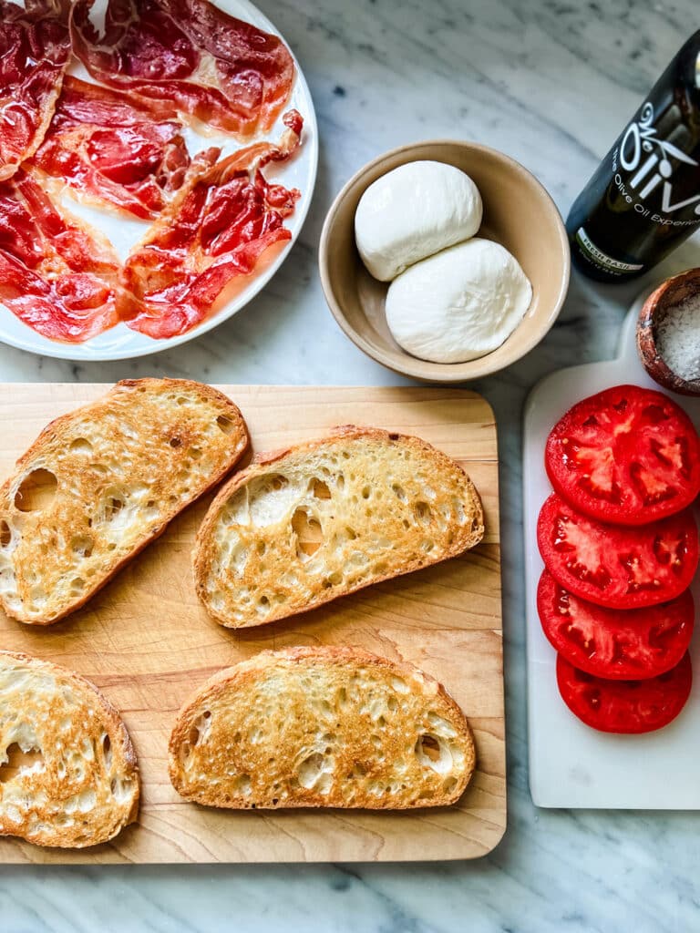 Toasted bread on a wooden cutting board is surrounded by fried prosciutto, a bowl of burrata and freshly sliced tomatoes right off the vine.
