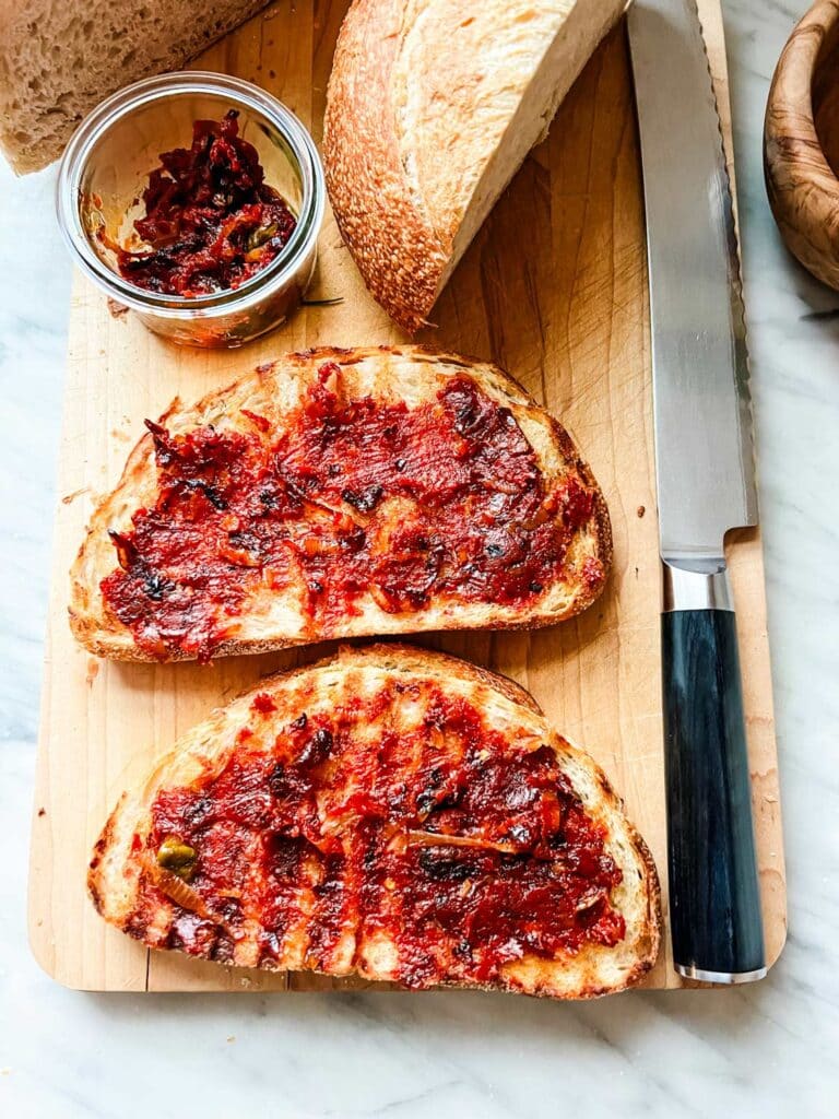 Toasted bread has shallot mixture spread on it sitting on a wooden cutting board. Extra spread is in a clear glass jar.