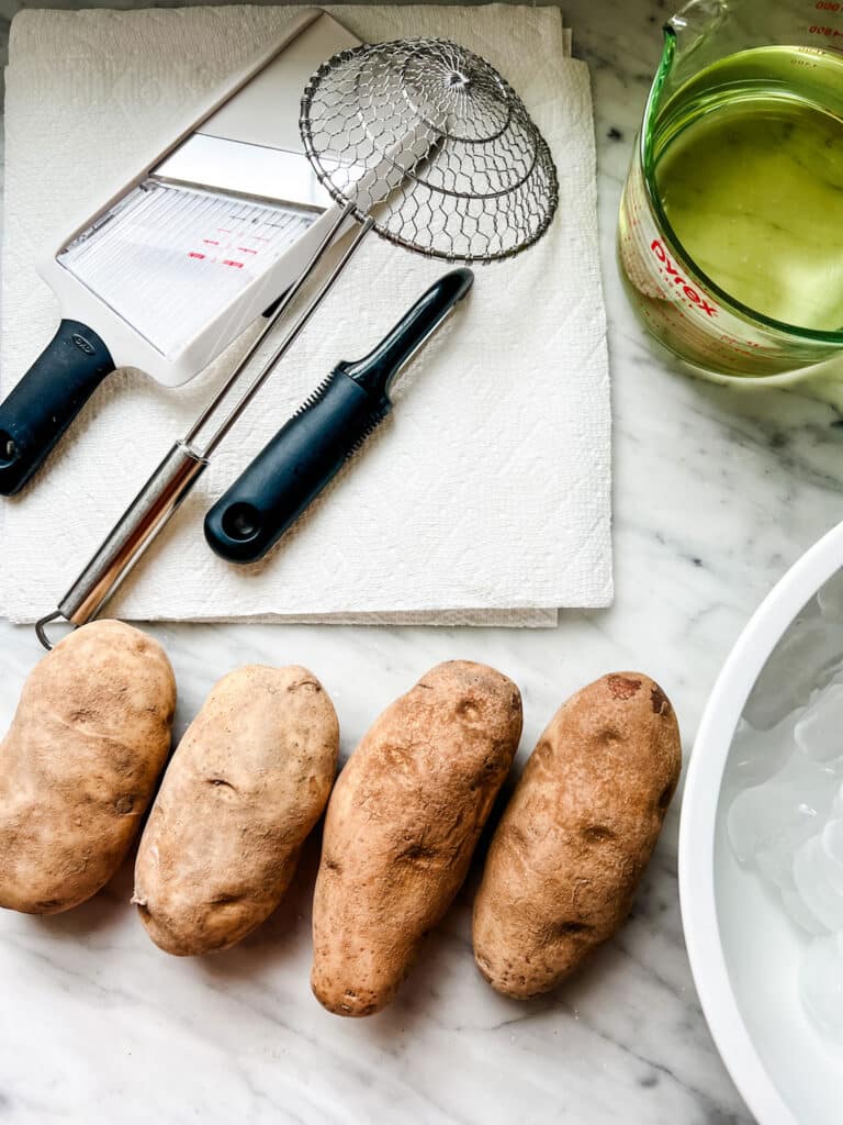 Four potatoes on a countertop with oil for frying, potato peeler, mandoline slicer, and spider drainer.