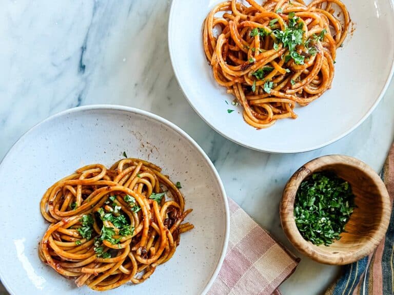 Caramelized shallot pasta is served in two white bowls. Next to the bowls is chopped parsley and garlic for garnishing.