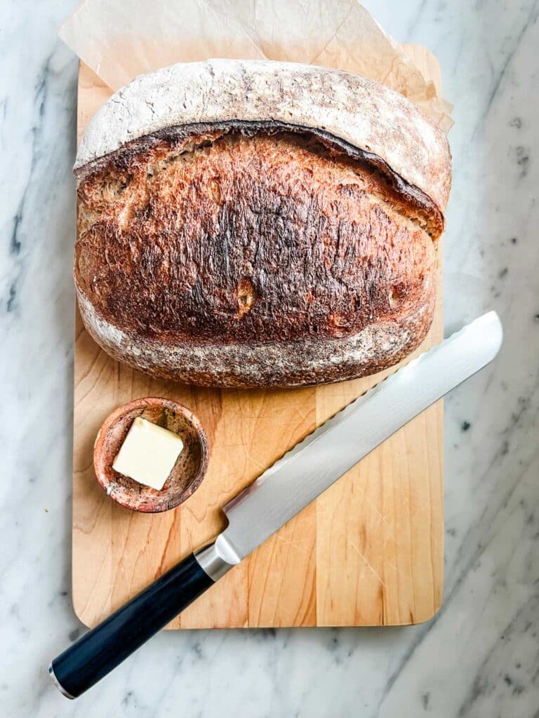 A loaf of rustic whole-grain bread is on a wooden cutting board with butter in a small stone bowl next to a Quince serrated bread knife.