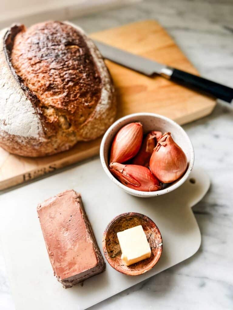 Unpeeled shallots are in a small white bowl next to a terrine of paté, butter and a loaf of rustic whole-grain bread.