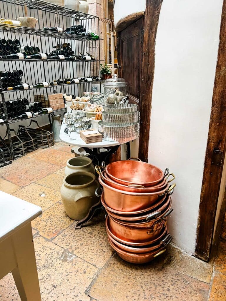 A tall stack of copper preserving pans on the floor in The Cook's Atelier. In the background, the shelves are stocked with all kinds of cooking products.