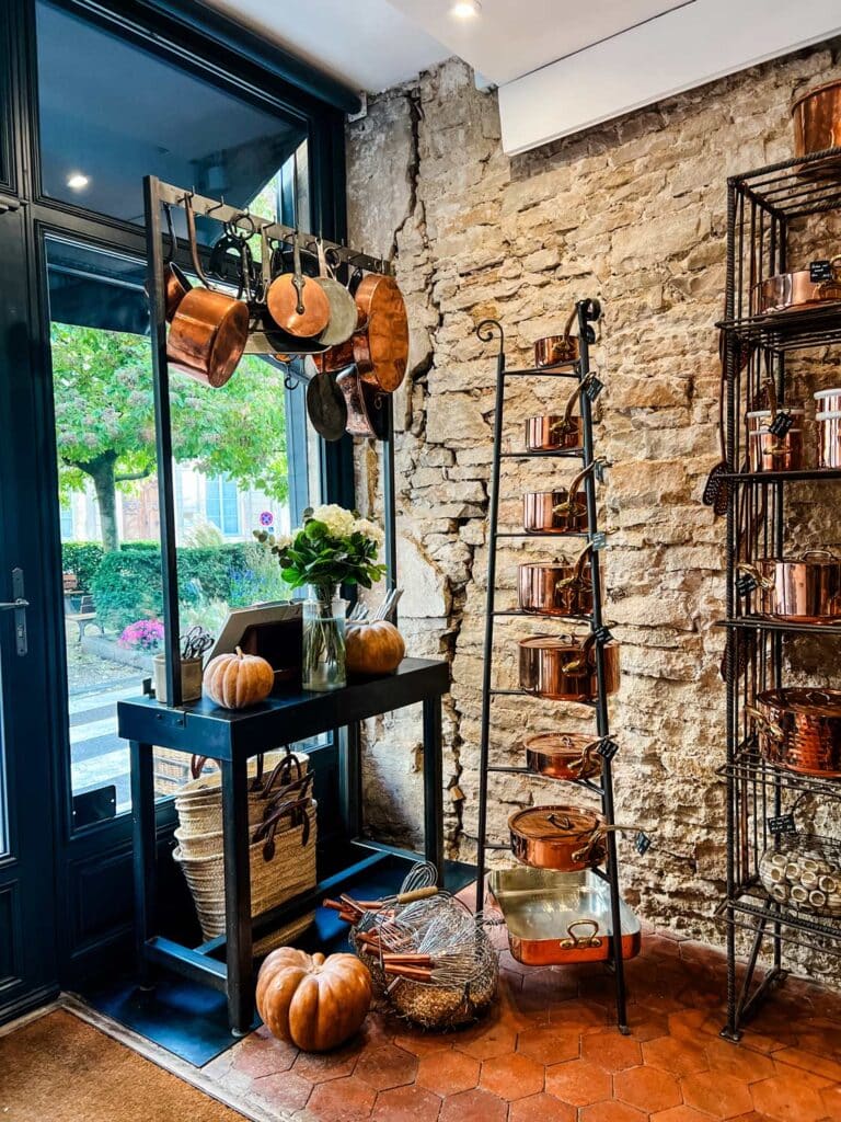 A kitchen island with hanging copper pots is featured in the window of The Cook's Atelier. A standing rack of all sizes of copper pots is next to it. 