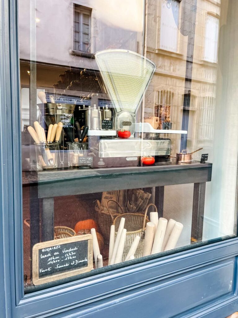 A close-up photo of the front window of The Cook's Atelier features a large cooking scale, knives, rolling pins and small copper pots.