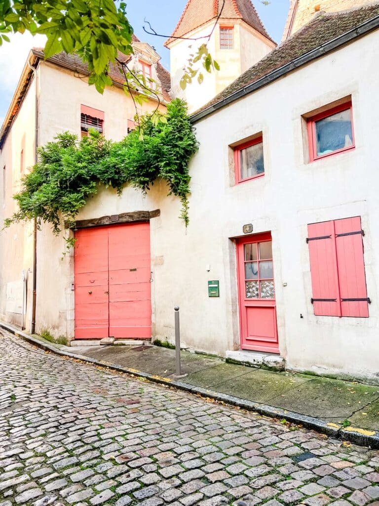 A small apartment on a cobblestone street has pink garage doors, a. pink front door, pink shutters, and pink window frames. Green moss is growing on the sidewalk.