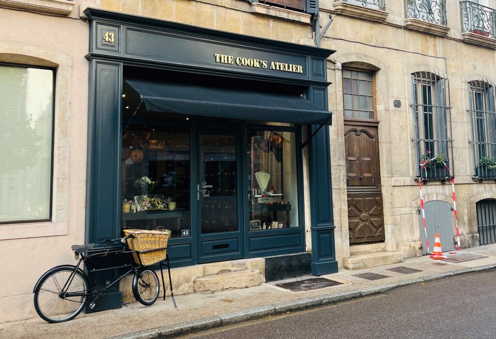 The Cook's Atelier, a cooking store in Beaune, France, has a beautiful exterior. An old-fashioned bicycle with a large basket on the front is parked in front of the shop.