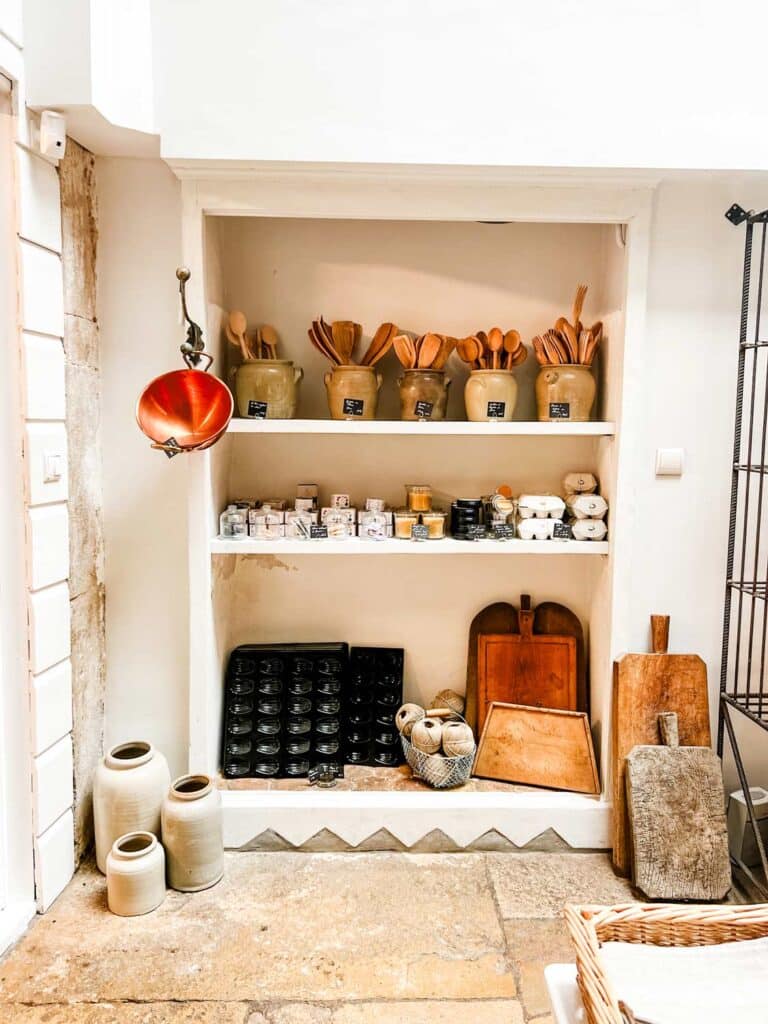 Vintage crocks full of wooden spoons line the top shelf of a display. On the second shelf is an assortment of spread and accouterments. On the third shelf of Madeleine tins, twine, and an assortment of wooden cutting boards. A copper pot hangs from one corner.