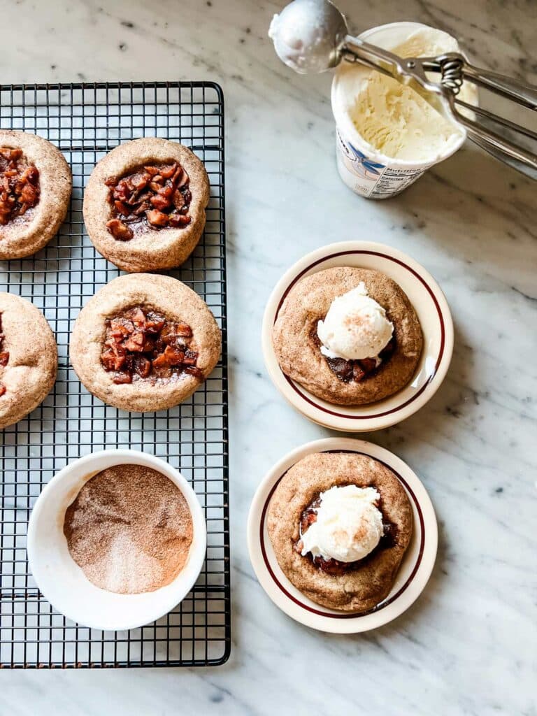 Two apple pie thumbprint cookies are on small plates and topped with a scoop of vanilla ice cream. Next to the plates on a wire cooking rack are the remaining cookies. A small bowl with cinnamon sugar is also on the cooling rack.